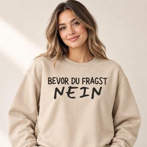 Op de afbeelding: Een beige sweater met ronde hals en de zwarte tekst BEVOR DU FRAGST NEIN in het midden op de borst gedrukt. Het kledingstuk heeft lange mouwen, een geribbelde halslijn en geribbelde boorden voor een standaard pasvorm.