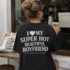 Könnte beinhalten: Ein schwarzes kurzärmeliges T-Shirt mit weißem Text auf der Rückseite, der I LOVE MY SUPER HOT BEAUTIFUL BOYFRIEND SO PLEASE STAY AWAY FROM ME lautet. Das T-Shirt hat einen Rundhalsausschnitt und eine lockere Passform, wobei die Grafik deutlich sichtbar ist.