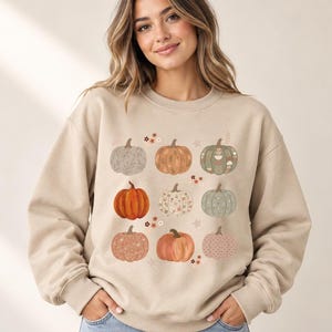 Peut inclure: Un sweat-shirt à col rond beige orné d'un imprimé graphique représentant neuf citrouilles disposées en grille. Les citrouilles sont illustrées dans des tons orange, vert et gris, chacune décorée de motifs uniques comme des points et des fleurs.
