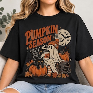 Op de afbeelding: Een zwart t-shirt met een grafische print met de tekst PUMPKIN SEASON, GOOD COFFEE SPOOKY VIBES, PUMPKIN SPICE, GHOST STORIES, FALL VIBES en WITCHY THINGS. Het ontwerp toont een geest met een muts die een koffiebeker vasthoudt, een zwarte kat, pompoenen, boeken en een maan met vleermuizen.