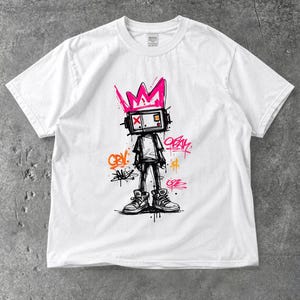 Op de afbeelding: Een wit t-shirt met een grafische print van een personage met een televisie als hoofd, dat een roze kroon draagt. Het ontwerp bevat graffiti-achtige tekstelementen in roze en oranje, met zwarte omlijningen en details van inktspatten.