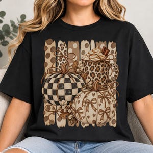 Pode incluir: Uma t-shirt preta com uma estampa gráfica de duas abóboras e uma caneca. Uma abóbora tem um padrão xadrez preto e branco, enquanto a outra é creme com laços e corações castanhos. A caneca tem estampa de leopardo e contém chantilly.