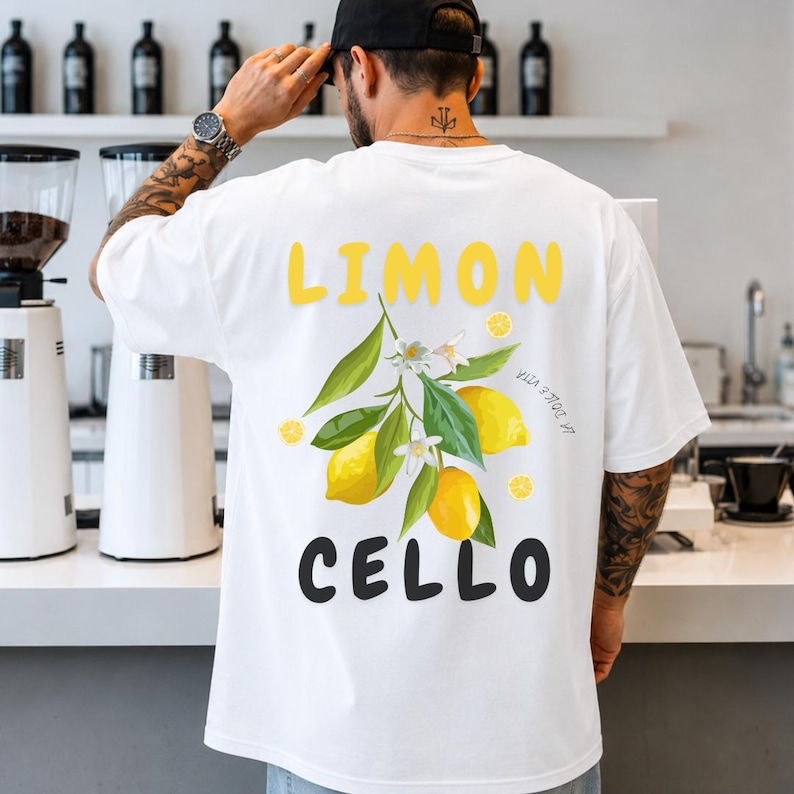 K&ouml;nnte beinhalten: Ein wei&szlig;es Kurzarm-T-Shirt mit einer Grafik auf der R&uuml;ckseite, die den gelben Schriftzug LIMON &uuml;ber einem B&uuml;ndel gr&uuml;ner Zitronenbl&auml;tter, wei&szlig;en Bl&uuml;ten und gelben Zitronen zeigt, sowie dem schwarzen Schriftzug CELLO und LA DOLCE VITA.