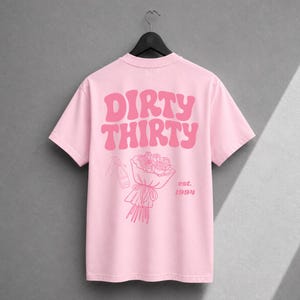 May include: A light pink t-shirt featuring the text DIRTY THIRTY in a bold, wavy font. Below the text is a line art illustration of a bouquet of roses and a champagne bottle, with the text est. 1994 printed to the right of the bouquet.