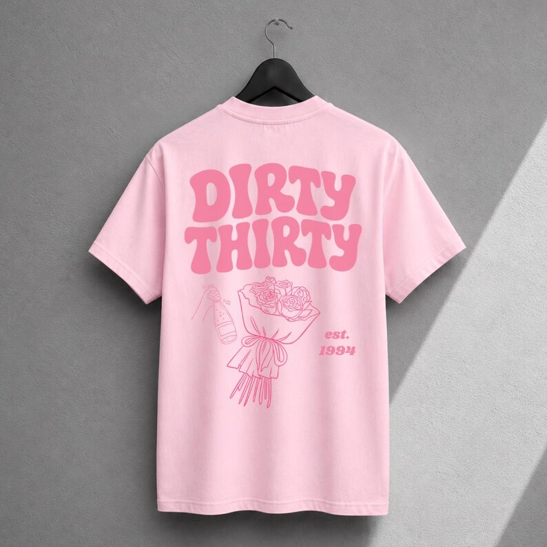 May include: A light pink t-shirt featuring the text DIRTY THIRTY in a bold, wavy font. Below the text is a line art illustration of a bouquet of roses and a champagne bottle, with the text est. 1994 printed to the right of the bouquet.
