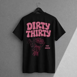 May include: A black short-sleeve t-shirt featuring pink graphic text that reads DIRTY THIRTY. Below the text is a line art illustration of a bouquet of roses and a champagne bottle, with the text est. 1994 printed to the right of the bouquet.