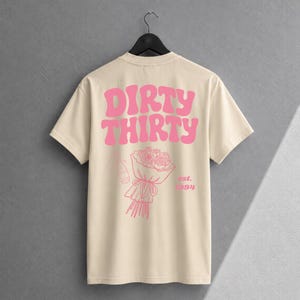 May include: A cream-colored short-sleeve t-shirt featuring pink graphic text that reads DIRTY THIRTY. Below the text is a line-art illustration of a bouquet of roses and a bottle, with the pink text est. 1994 printed to the right of the bouquet.