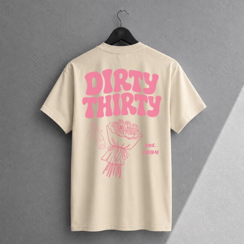 May include: A cream-colored short-sleeve t-shirt featuring pink graphic text that reads DIRTY THIRTY. Below the text is a line-art illustration of a bouquet of roses and a bottle, with the pink text est. 1994 printed to the right of the bouquet.