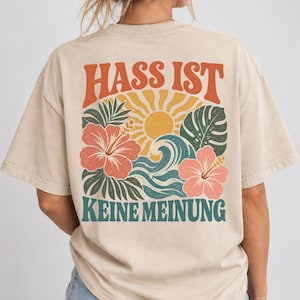 Könnte beinhalten: Ein beiges Kurzarm-T-Shirt mit einem Grafikdruck auf der Rückseite, der den Text HASS IST KEINE MEINUNG in Orange und Petrol zeigt. Das Motiv umfasst eine gelbe Sonne, türkisfarbene Wellen sowie rosa Hibiskusblüten mit grünen Blättern.