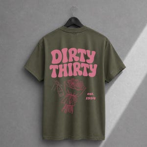 May include: An olive green short-sleeve t-shirt featuring the pink text DIRTY THIRTY in a wavy font. Below the text is a pink line-art illustration of a hand holding a champagne bottle and a bouquet of roses, with the pink text est. 1994.