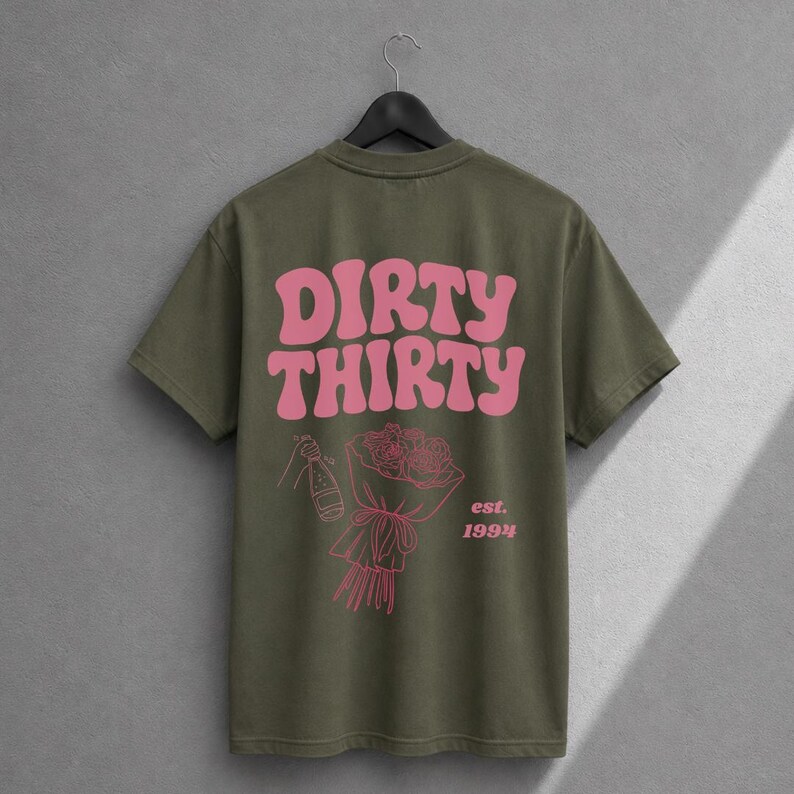 May include: An olive green short-sleeve t-shirt featuring the pink text DIRTY THIRTY in a wavy font. Below the text is a pink line-art illustration of a hand holding a champagne bottle and a bouquet of roses, with the pink text est. 1994.