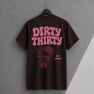 May include: A dark brown short-sleeve t-shirt featuring the pink text DIRTY THIRTY in a wavy font. Below the text is a pink line-art graphic of a hand holding a champagne bottle next to a bouquet of roses with the text est. 1994 printed in pink.