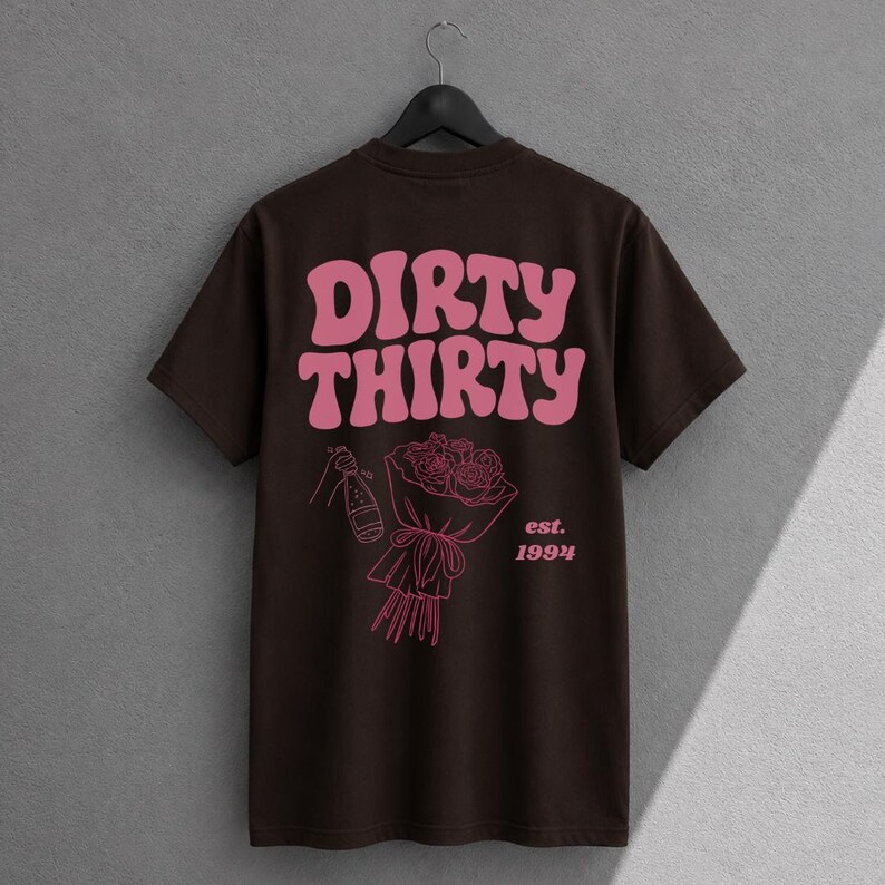 May include: A dark brown short-sleeve t-shirt featuring the pink text DIRTY THIRTY in a wavy font. Below the text is a pink line-art graphic of a hand holding a champagne bottle next to a bouquet of roses with the text est. 1994 printed in pink.