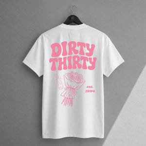 May include: A white short-sleeve t-shirt featuring pink graphic text that reads DIRTY THIRTY. Below the text is a line art illustration of a bouquet of roses and a champagne bottle, with the text est. 1994 printed to the right of the bouquet.