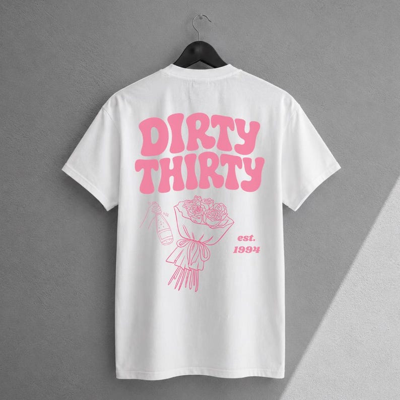 May include: A white short-sleeve t-shirt featuring pink graphic text that reads DIRTY THIRTY. Below the text is a line art illustration of a bouquet of roses and a champagne bottle, with the text est. 1994 printed to the right of the bouquet.