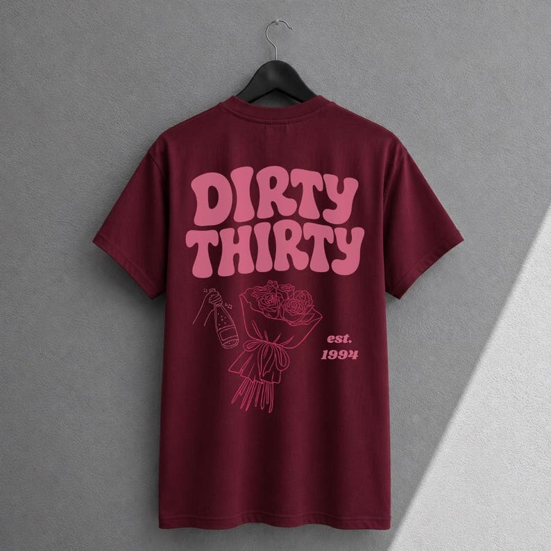 May include: A maroon short-sleeve t-shirt featuring a pink graphic on the back. The design includes the text "DIRTY THIRTY" in a wavy font, a line drawing of a champagne bottle, a bouquet of roses, and the text "est. 1994" printed below.