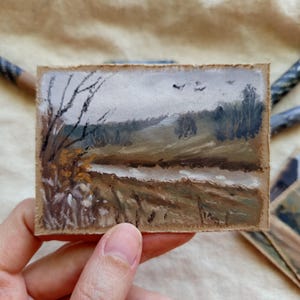 Può includere: Piccolo dipinto di paesaggio su carta ruvida. L'opera raffigura una scena all'aperto serena con un fiume, alberi e un cielo nuvoloso. La tavolozza dei colori comprende tonalità tenui di marrone, verde e grigio.
