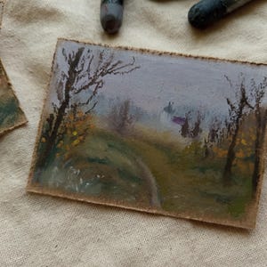 May include: Small landscape painting on a rectangular piece of paper. The artwork depicts a path winding through a misty forest, with trees and foliage in muted greens, yellows, and browns. The sky is a soft lavender.