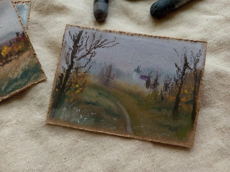 May include: Small landscape painting on a rectangular piece of paper. The artwork depicts a path winding through a misty forest, with trees and foliage in muted greens, yellows, and browns. The sky is a soft lavender.