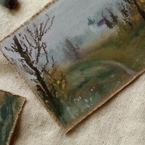 May include: Oil pastel landscape painting on textured paper. The artwork depicts a path winding through a misty forest, with a bare tree in the foreground. The color palette includes muted greens, browns, and grays, with touches of yellow.