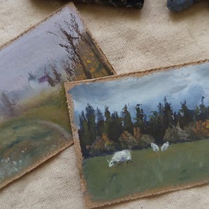 May include: Two small landscape paintings on textured paper. One depicts a misty path with trees, the other shows sheep grazing in a field near a forest under a cloudy sky. The paintings have a rustic, handmade feel.