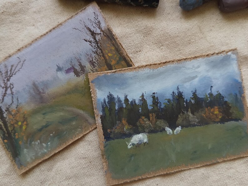 May include: Two small landscape paintings on textured paper. One depicts a misty path with trees, the other shows sheep grazing in a field near a forest under a cloudy sky. The paintings have a rustic, handmade feel.