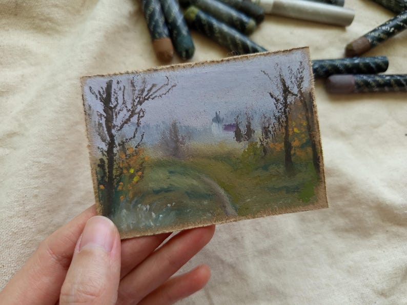 May include: A small, rectangular landscape painting featuring a path winding through a misty forest. The artwork uses pastel colors, with a focus on greens, yellows, and purples. Bare trees and a distant building are visible.