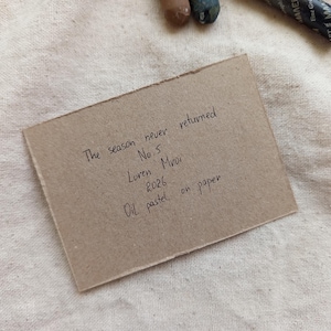 May include: A small, rectangular piece of brown paper with handwritten text, including "The season never returned" and "Oil pastel on paper." The paper rests on a textured, off-white fabric, with art supplies visible.