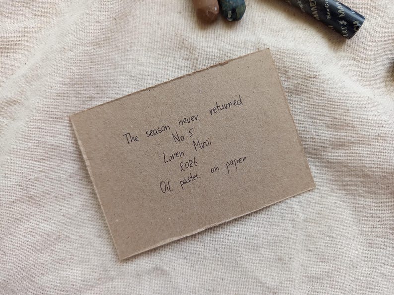 May include: A small, rectangular piece of brown paper with handwritten text, including "The season never returned" and "Oil pastel on paper." The paper rests on a textured, off-white fabric, with art supplies visible.