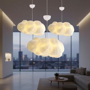 May include: Three cloud-shaped pendant lights hanging from the ceiling. The lights are white with a soft, textured appearance, and they emit a warm, inviting glow. The lights are suspended in a modern living room with a city view.