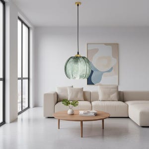 May include: A hanging pendant light with a green, ribbed glass shade and a gold-colored canopy. The light fixture hangs from a black cord and is suspended above a round wooden coffee table. The room has a modern, minimalist design.