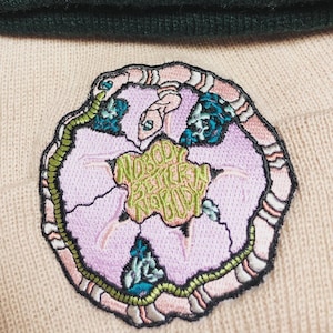 May include: A pink and green embroidered patch with a flower design and the text "Nobody Better Than Nobody".