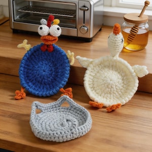 May include: A collection of handmade crochet trivets in the shape of a chicken, a duck, and a cat. The chicken trivet is blue with orange and yellow accents. The duck trivet is white with orange accents. The cat trivet is gray.