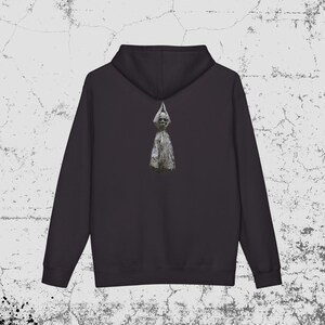 Puede incluir: Una sudadera con capucha negra que presenta un gráfico centrado en la parte posterior. El gráfico muestra una figura pálida similar a una calavera envuelta en una tela texturizada de color claro que cuelga hacia abajo, formando una punta.