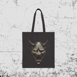 May include: A black tote bag featuring a centered graphic of a weathered, horned Hannya mask with a single visible white eye and dark markings. Below the mask, the word ZANGAI is printed in small white capital letters on the fabric surface.