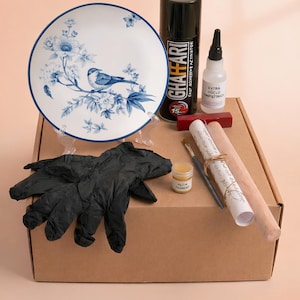 Può includere: Un kit artigianale con un piatto bianco con un design di uccelli e fiori blu. Il kit include colla, vernice, pennelli, guanti e tasselli di legno, il tutto all'interno di una scatola di cartone.