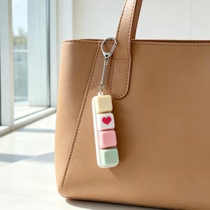 May include: A pastel-colored keychain with four square blocks, attached to a tan handbag. The top block is yellow, the second has a red heart, the third is pink, and the bottom is mint green. The keychain is attached with a silver clasp.