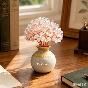 May include: A decorative ornament featuring a small, round, white and tan ceramic vase with the text To Grace printed on the front. Inside the vase is a wire tree sculpture with numerous small, translucent, pale pink crystal beads.