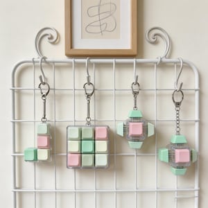 Puede incluir: Cuatro llaveros con diseños cuadrados en colores pastel se exhiben en una rejilla de alambre blanca. Los llaveros presentan cuadrados rosas, verdes y blancos, algunos dispuestos en forma de cubo, otros en diseño de cruz. Cada llavero tiene un cierre plateado.