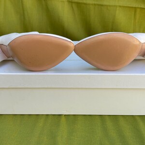 May include: Pair of white leather high heels with a pointed toe and a small, square heel. The soles are a light tan color. The shoes are resting on a white box, with a green background.