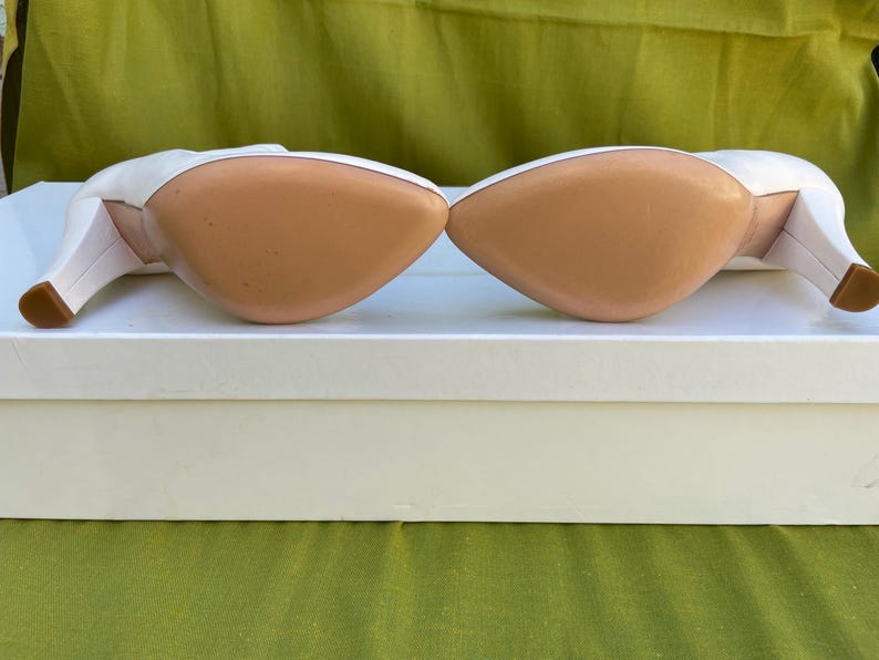 May include: Pair of white leather high heels with a pointed toe and a small, square heel. The soles are a light tan color. The shoes are resting on a white box, with a green background.