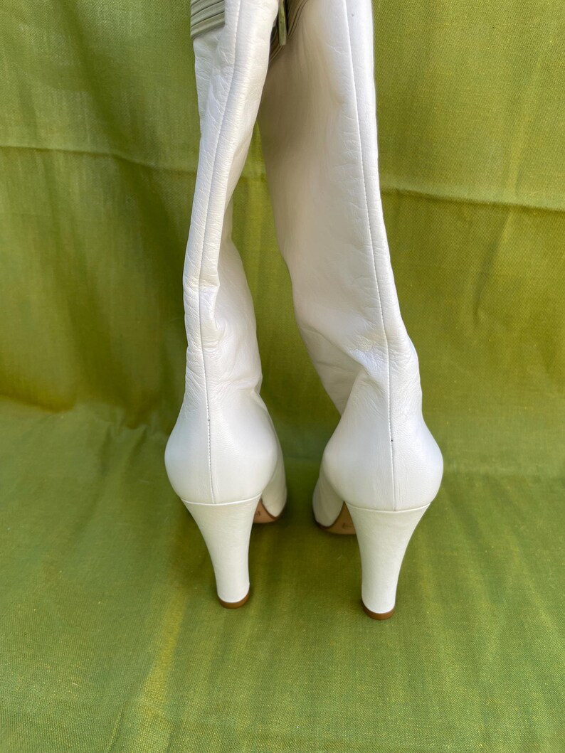 May include: A pair of white leather knee-high boots with a pointed toe and a curved heel. The boots are set against a green background, showcasing their sleek design and elegant silhouette.