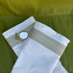 May include: A pair of white leather gloves with a light beige band at the wrist. A small, round mirror and a gold triangle are attached to one glove. The gloves are set against a green background.