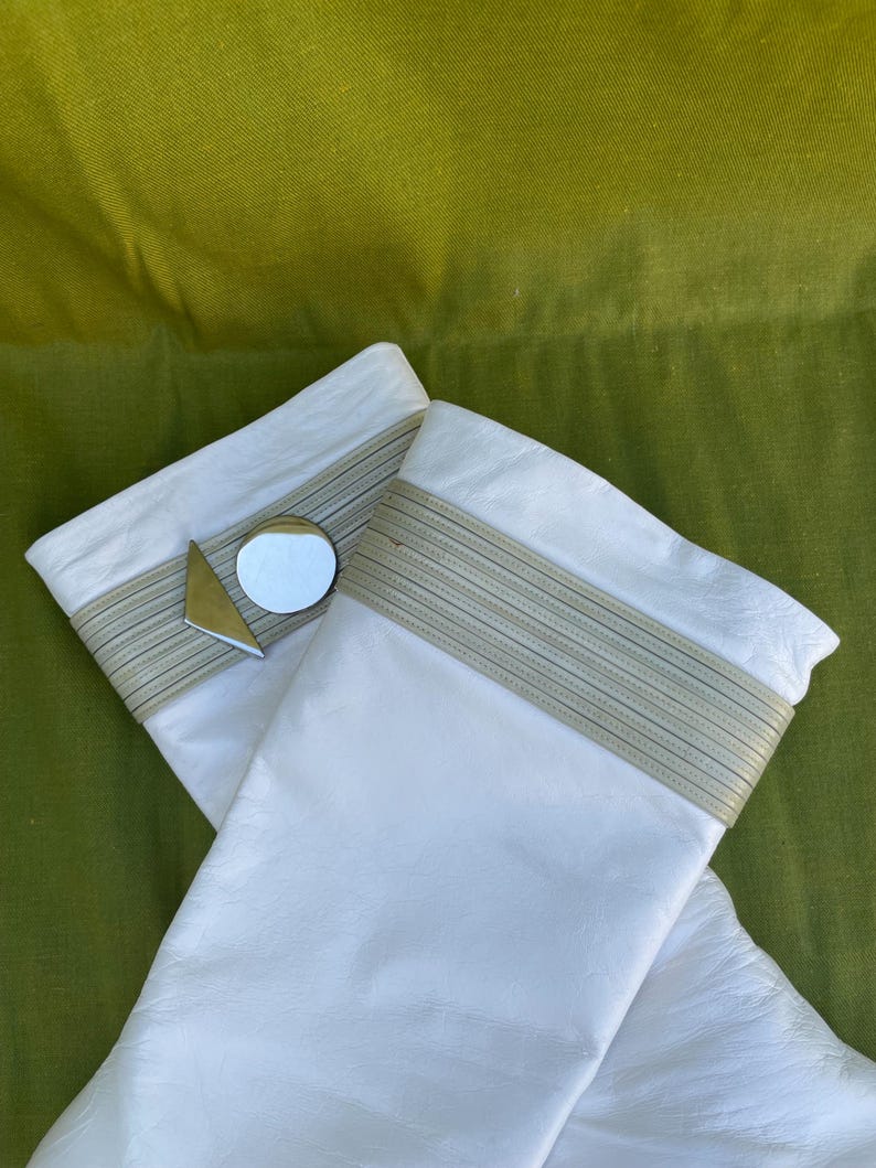 May include: A pair of white leather gloves with a light beige band at the wrist. A small, round mirror and a gold triangle are attached to one glove. The gloves are set against a green background.