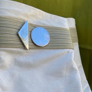 May include: Close-up of a white leather bag with a silver geometric clasp. The clasp features a diamond shape and a circle. The bag has a light gray band across the middle.