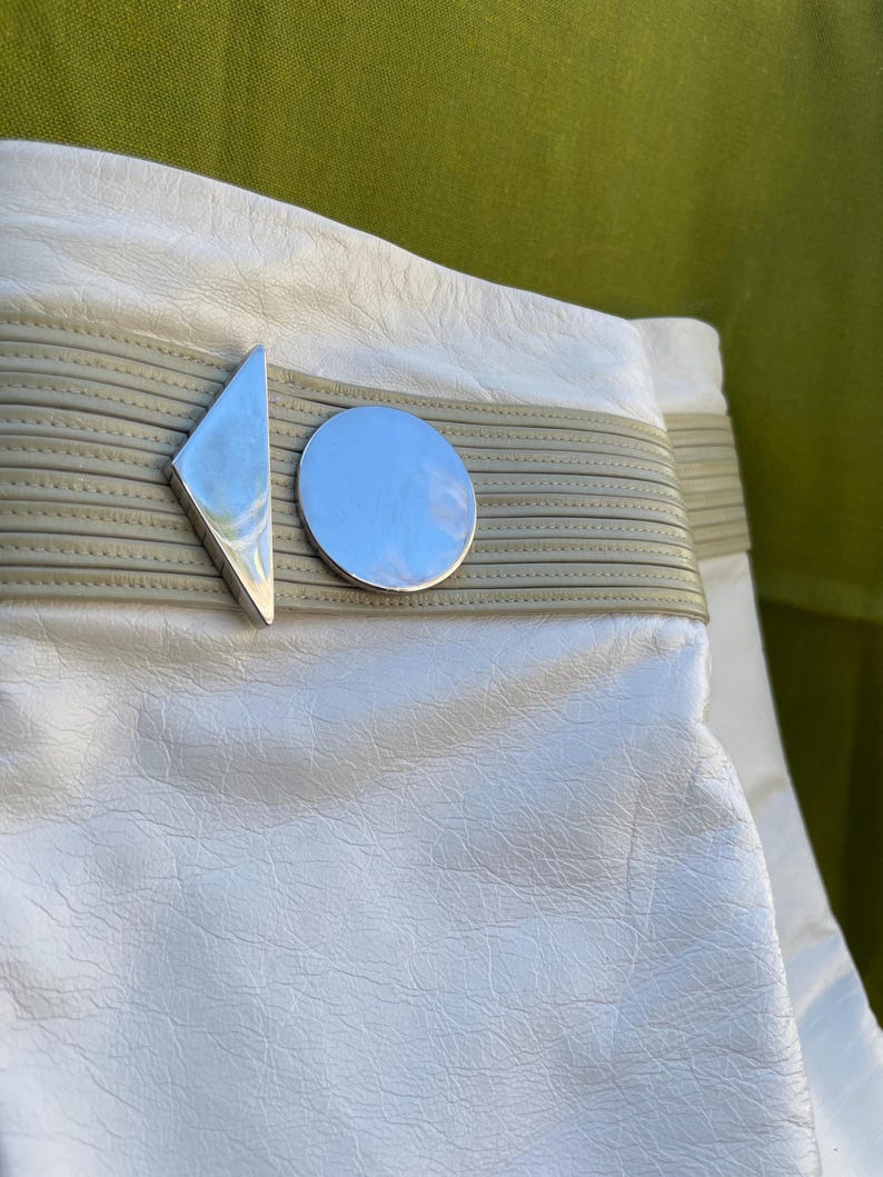 May include: Close-up of a white leather bag with a silver geometric clasp. The clasp features a diamond shape and a circle. The bag has a light gray band across the middle.