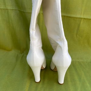 May include: A pair of white leather knee-high boots with a pointed toe and a curved heel. The boots are set against a green background, showcasing their sleek design and elegant silhouette.