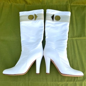 May include: A pair of white knee-high boots with a pointed toe and a stiletto heel. The boots have a decorative band at the top with a gold-colored circular accent and a small arrow-shaped detail. The soles are a light brown color.