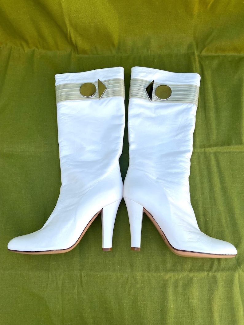 May include: A pair of white knee-high boots with a pointed toe and a stiletto heel. The boots have a decorative band at the top with a gold-colored circular accent and a small arrow-shaped detail. The soles are a light brown color.