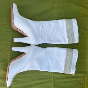 May include: A pair of white leather knee-high boots with a pointed toe and a stiletto heel. The boots have a beige sole and a beige elastic band detail at the top of the boot. The boots are set against a green background.